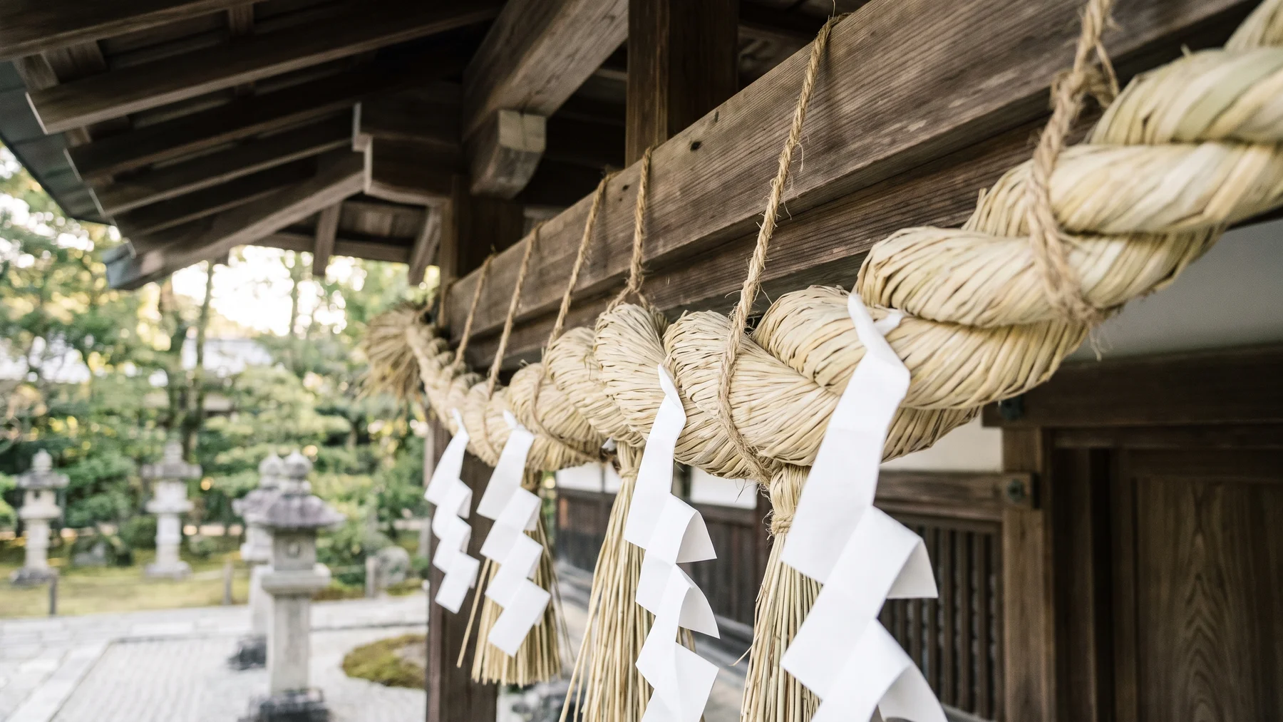 Imagined close-up of a straw shimenawa rope and folded white shide under wooden shrine eaves.