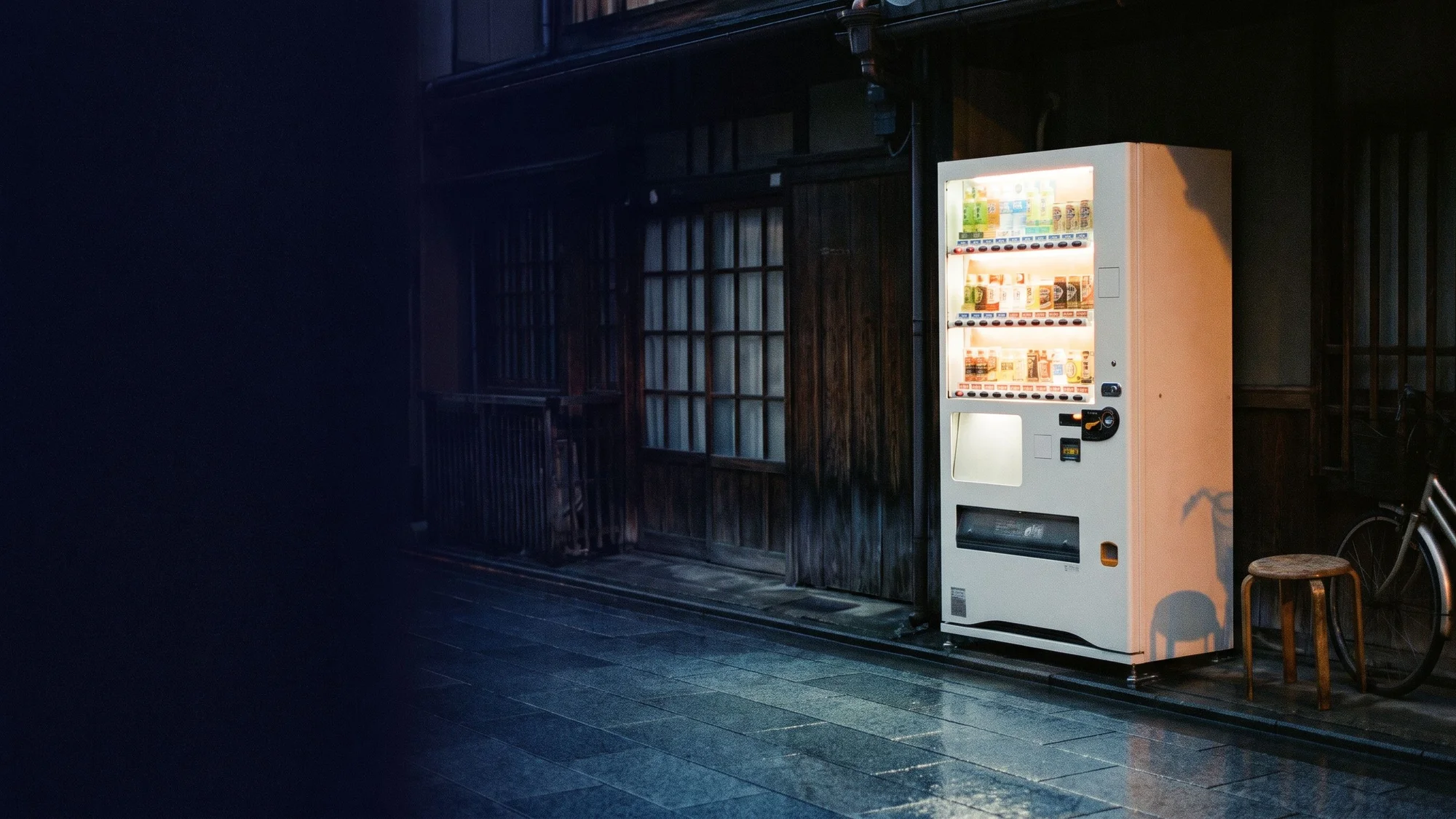 A glowing beverage machine outside a wooden storefront on a wet, dark street.